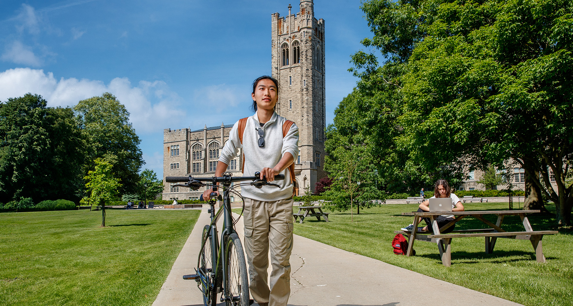 A person walks with a bicycle on a sunny university campus. A historic stone building is in the background, while another person uses a laptop on a picnic table nearby.