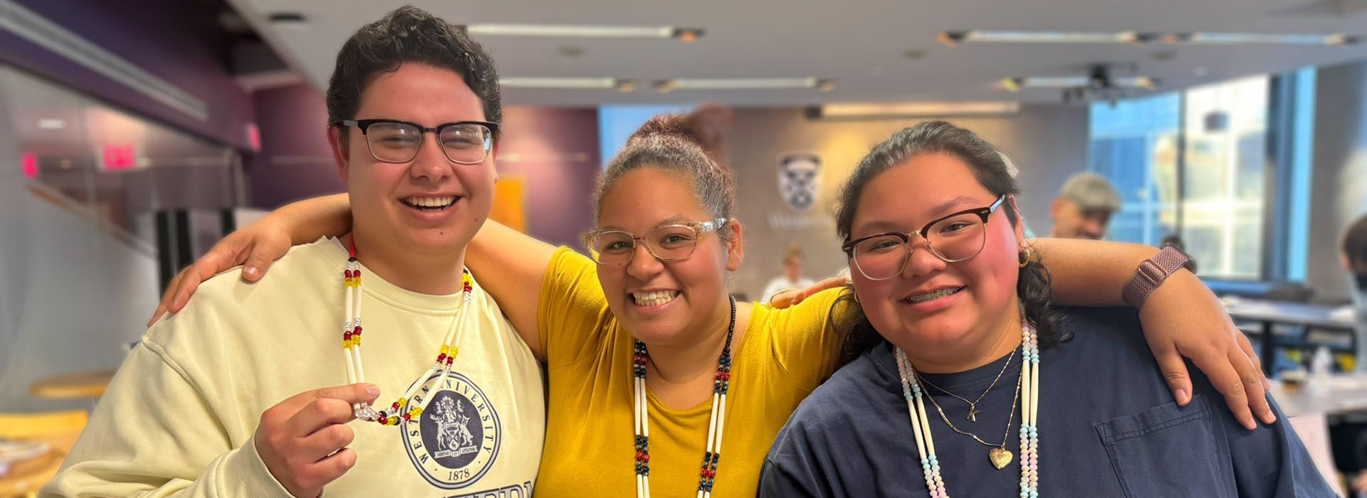 Three people standing indoors, smiling at the camera and wearing beaded necklaces, with one holding up their necklace